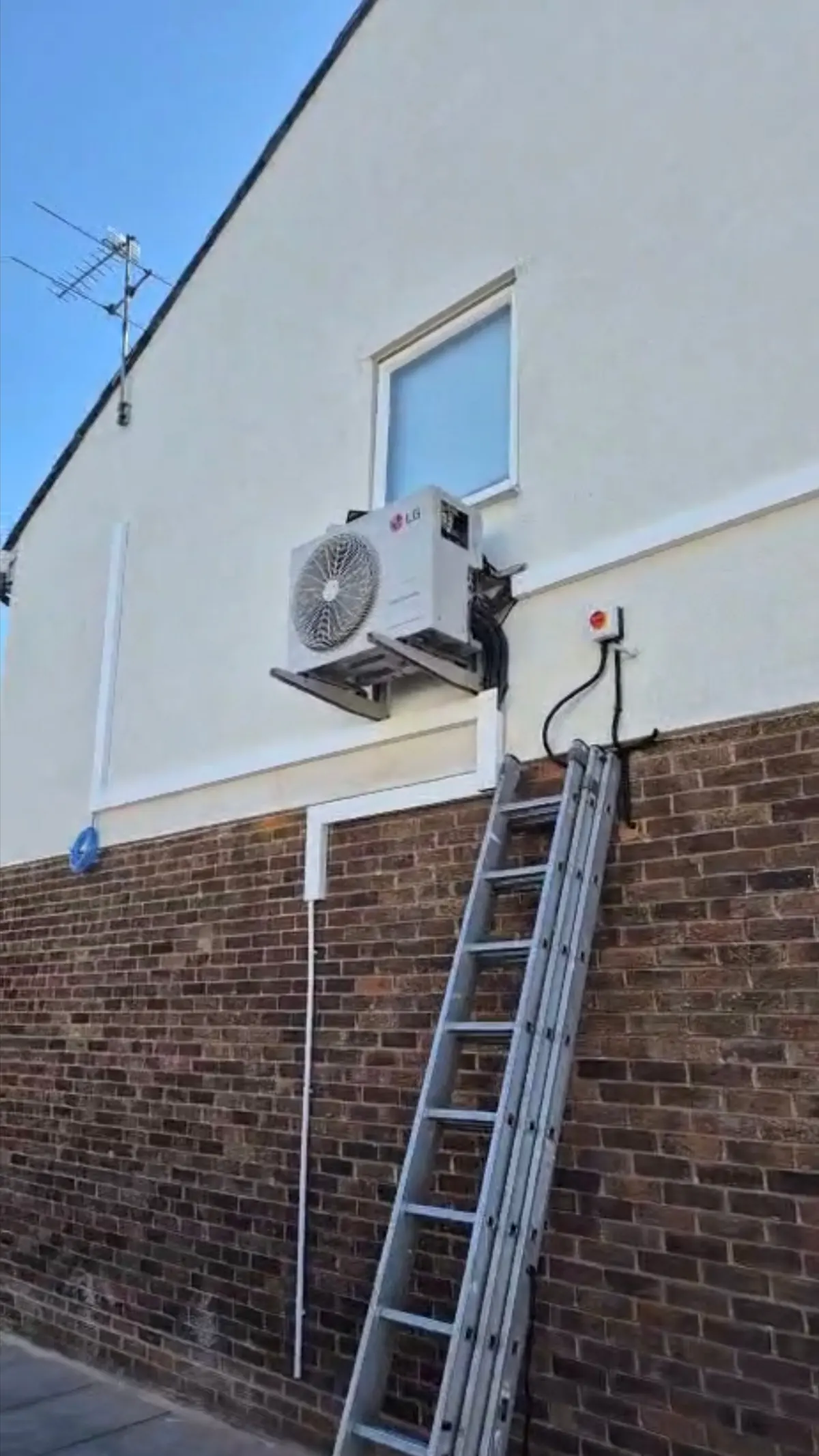 Air conditioning outdoor unit on brackets high on a rendered wall, with a ladder in position for access